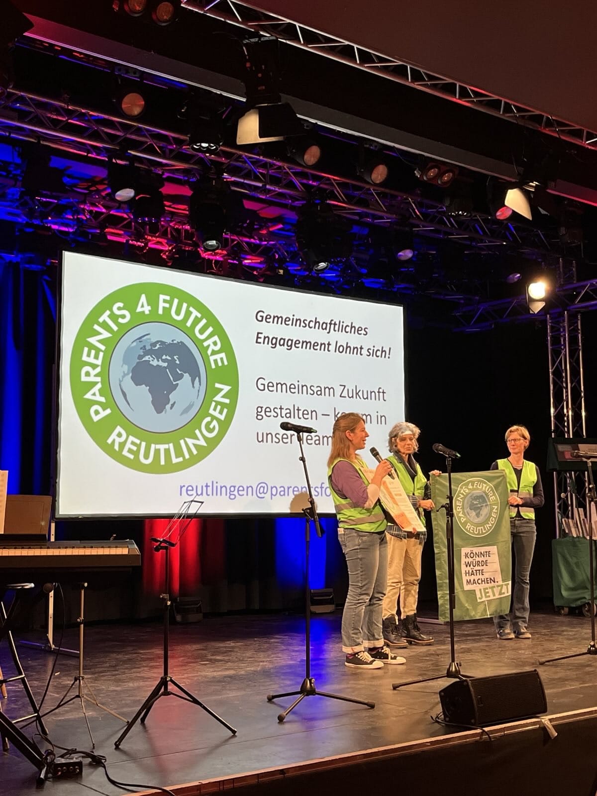 Drei Mitglieder der Parents for Future Reutlingen stehen auf einer Bühne. Sie tragen grüne Warnwesten. Zwei halten ein Banner mit dem Logo. Die dritte Person spricht in ein Mikrofon. Im Hintergrund sieht man eine Folie, auf der Links das Logo der Ortsgruppe ist, rechts stehen die Worte: Gemeinschaftliches Engagement lohnt sich! Gemeinsam Zukunft gestalten – komm in unser Team.